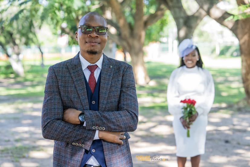 Confident groom portrait in checkered suit with bride in background