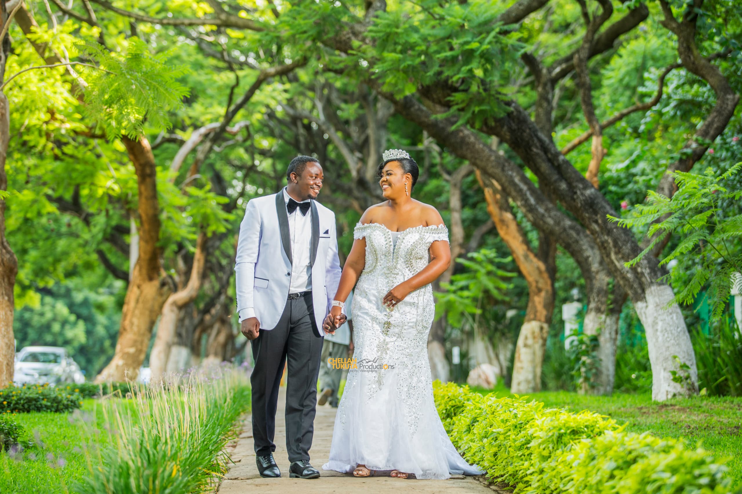Tree-lined romantic stroll - couple hand in hand, groom in white jacket, bride in sparkly gown